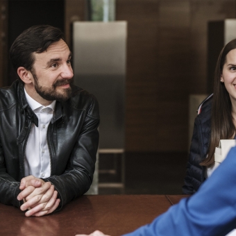 Zwei Personen stehen vor einem Bank Schalter und sprechen mit einem Mitarbeiter der Bank.